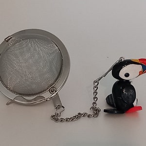May include: A silver tea infuser with a mesh ball and a chain attached to a small, colourful ceramic puffin figurine.