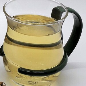 Puede incluir: Una taza de t&eacute; de vidrio transparente con un asa verde oscuro, llena de t&eacute; amarillo claro.