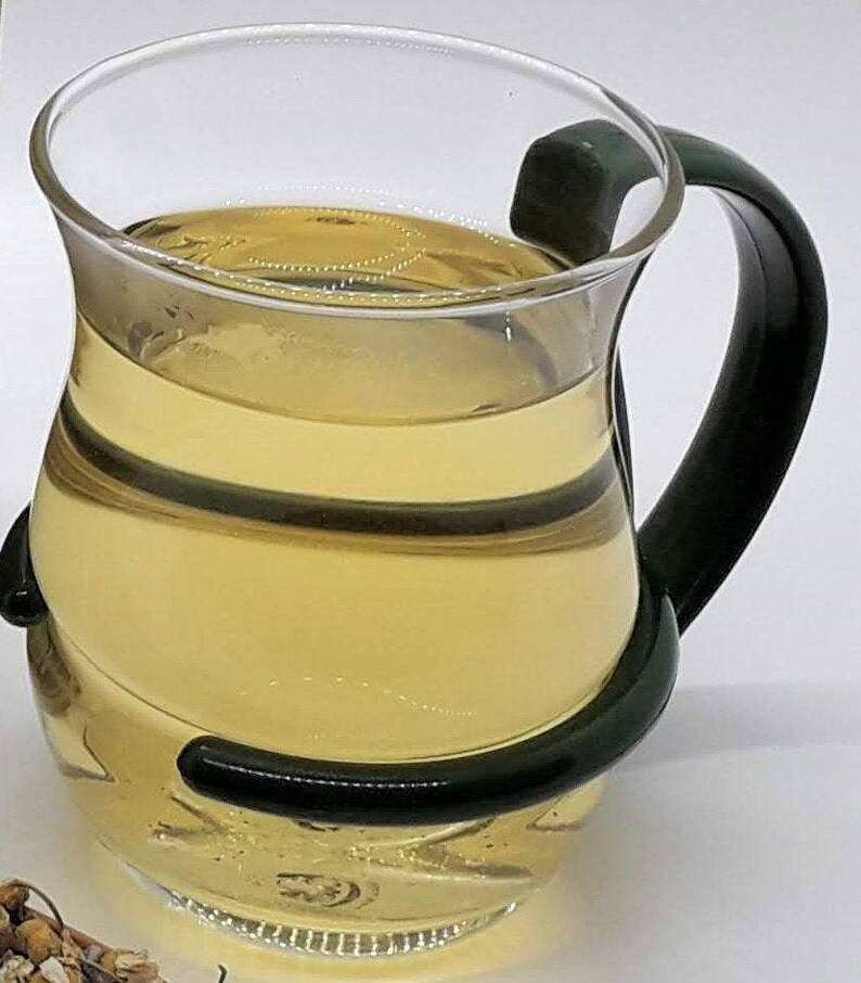 Puede incluir: Una taza de t&eacute; de vidrio transparente con un asa verde oscuro, llena de t&eacute; amarillo claro.