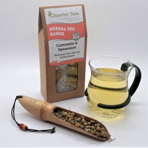 Pu&ograve; includere: Una scatola di cartone marrone con una finestra che mostra t&egrave; di camomilla e menta verde essiccato. La scatola ha un'etichetta che recita "Chantler Teas Herbal Tea Range Camomile & Spearmint Relaxing and calming herbal blend". Sono inoltre visibili una tazza di t&egrave; di vetro e una paletta di legno con foglie di t&egrave;.