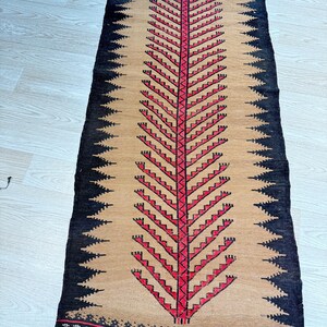 May include: A long, narrow rug with a beige background and a black and red tree design. The rug has a fringe on both ends.