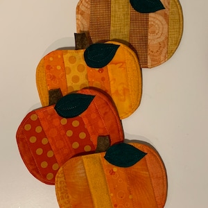 May include: Four orange and yellow fabric pumpkin-shaped coasters with green felt leaves. The coasters are made of different patterned fabrics and are perfect for adding a touch of fall decor to your home.