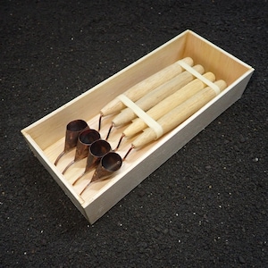 Peut inclure: Un ensemble de quatre outils tjanting avec des manches en bois clair et des réservoirs en cuivre, disposés dans une boîte en bois rectangulaire. Les outils sont maintenus ensemble par un élastique blanc, révélant leurs pointes métalliques.