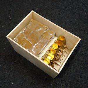 May include: A wooden box contains a wrapped package and several gold-colored objects. The objects have a cylindrical shape with a rounded top and a thin, pointed base. The box is made of light-colored wood.