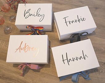 May include: Four white gift boxes with personalized names, "Bailey", "Frankie", "Aubrey", and "Hannah", are arranged on a wood floor. Each box is tied with a different colored ribbon.