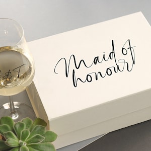 May include: A white gift box with the text "Maid of honour" in black script. A wine glass with the initials "A & T" is in the foreground.