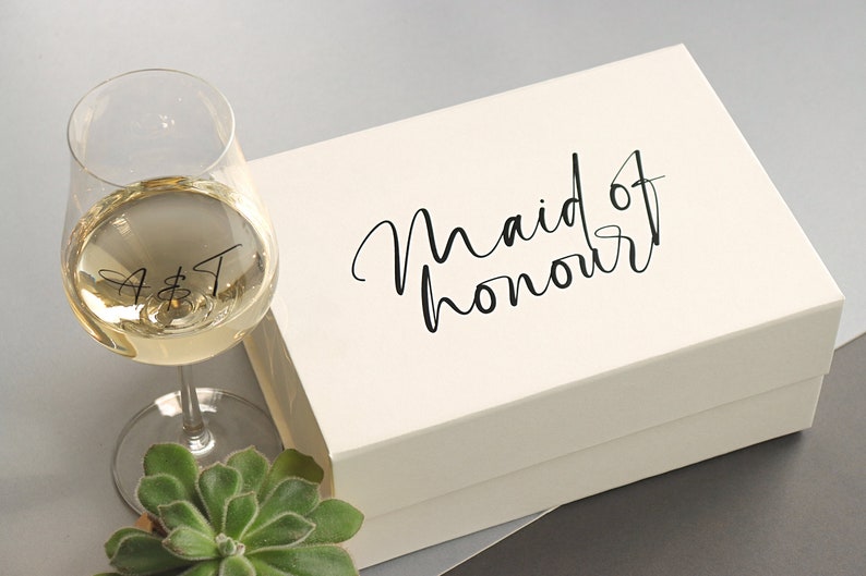 May include: A white gift box with the text "Maid of honour" in black script. A wine glass with the initials "A & T" is in the foreground.