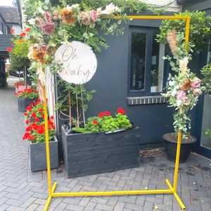 May include: A yellow metal archway decorated with dried flowers and greenery. The archway is set up outdoors in front of a building with a sign that reads "Oh baby!"