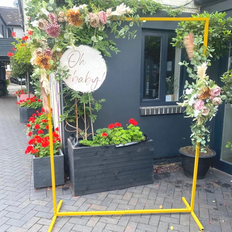 May include: A yellow metal archway decorated with dried flowers and greenery. The archway is set up outdoors in front of a building with a sign that reads "Oh baby!"