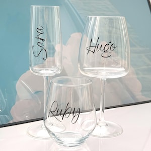 Puede incluir: Tres copas de cristal transparente: una flauta de champán con "Sara", una copa de vino con "Hugo" y un vaso sin pie con "Ruby" en escritura negra. Las copas están dispuestas sobre una superficie blanca con un fondo azul borroso.