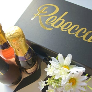 May include: A black gift box with the name "Rebecca" in gold lettering. The box is surrounded by three mini bottles of champagne, one pink and two clear, and a bouquet of white daisies.