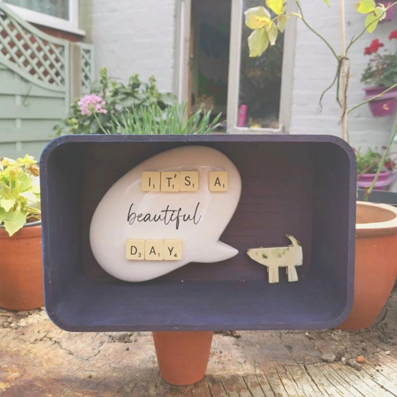 May include: A wooden box with a purple interior, featuring a white ceramic speech bubble with the words "It's a beautiful day" spelled out with letter tiles. A small ceramic dog figurine is also included.
