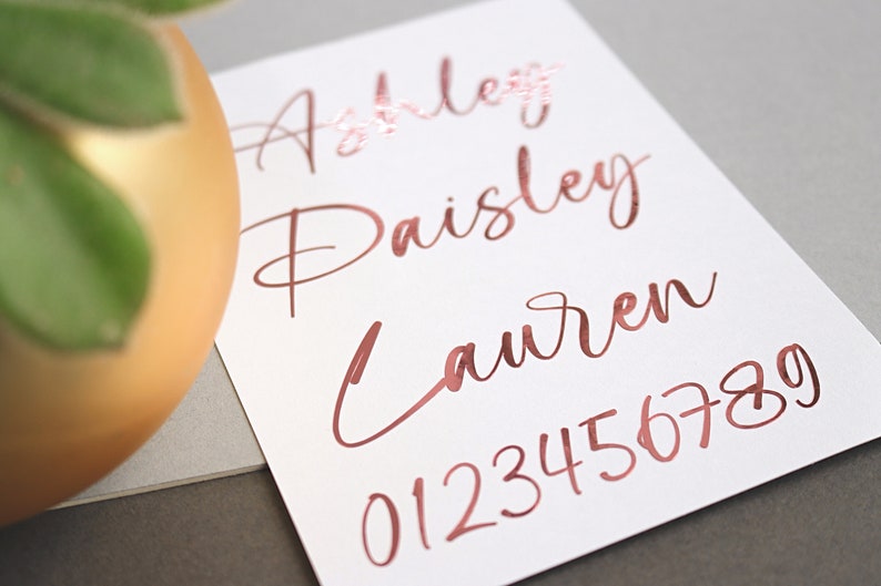 May include: A white card with the names Ashley, Paisly, and Lauren written in rose gold foil lettering. The numbers 0123456789 are also written in rose gold foil lettering.