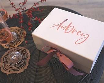May include: A white gift box with the name "Aubrey" in a copper-colored script, tied with a matching ribbon. Two champagne flutes with pink liquid and wooden coasters are also visible.