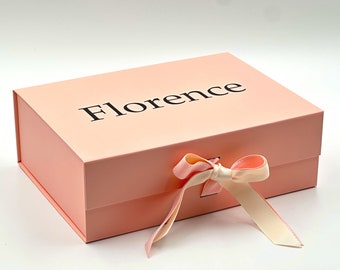 Op de afbeelding: Een rechthoekige lichtroze geschenkdoos met een scharnierend deksel. Het woord Florence is in een zwart schreeflettertype op de bovenkant gedrukt. Een crèmekleurig lint is in het midden aan de voorzijde gestrikt om het deksel te sluiten.
