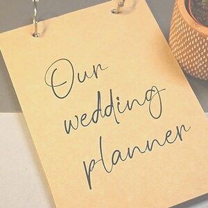 May include: A brown paper wedding planner with a silver ring binder and the text "Our wedding planner" written in black cursive.