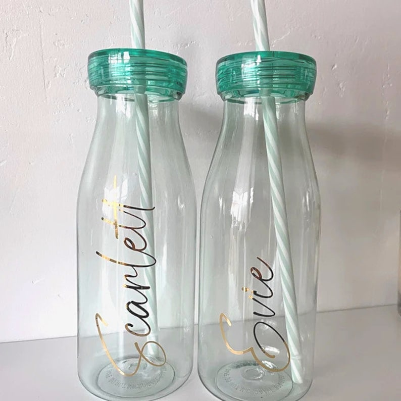 May include: Two clear glass water bottles with mint green lids and matching striped straws. The bottles have gold lettering that reads "Scarlett" and "Evie".