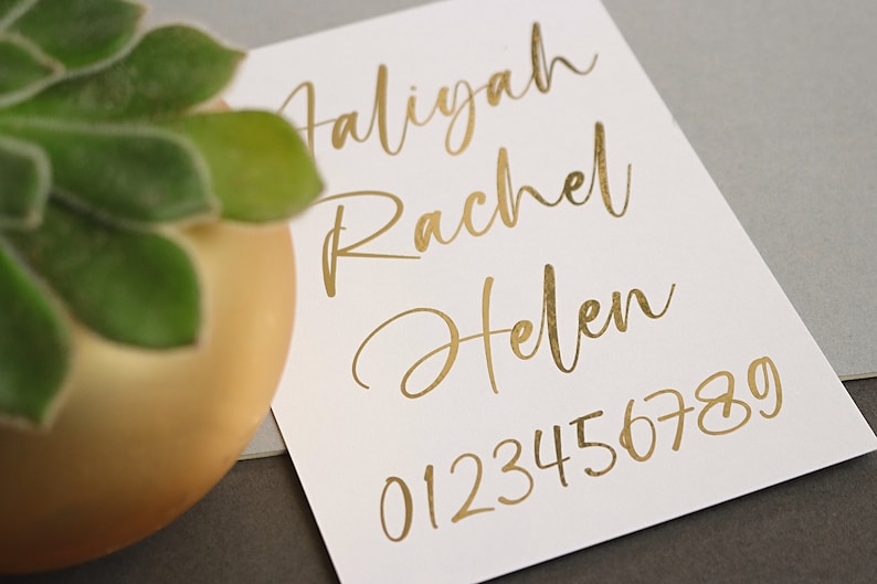May include: A white card with gold lettering. The names Aliyah, Rachel, and Helen are written in cursive. The numbers 0123456789 are written in a simple font.