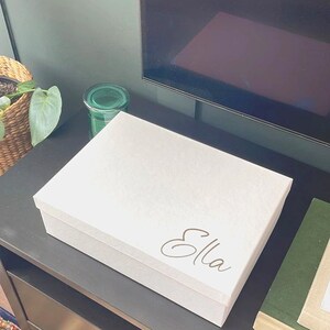 May include: White storage box with the name "Ella" written in gold cursive lettering.