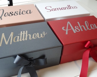 May include: Four gift boxes with personalized names on the top. The boxes are in different colors: pink, white, gray, and red. Each box has a ribbon tied around it.