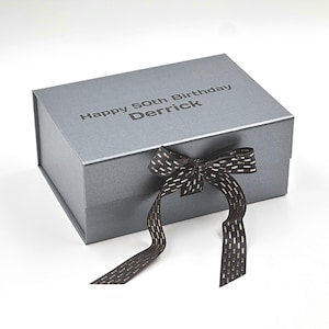 Pode incluir: Uma caixa de presente cinza com uma fita preta e detalhes dourados. A caixa é personalizada com o texto "Happy 50th Birthday Derrick".