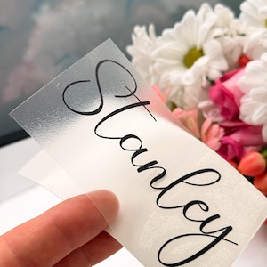 Könnte beinhalten: Ein klarer Vinyl-Aufkleber mit dem Namen "Stanley" in eleganter schwarzer Schrift. Der Aufkleber ist teilweise abgezogen, wodurch eine weiße Rückseite sichtbar wird. Im Hintergrund befindet sich ein Strauß weißer und rosa Blumen.