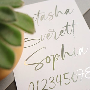 May include: A white card with the names "Natasha Everett Sophia" written in silver foil lettering. The numbers "0123456789" are also written in silver foil lettering.