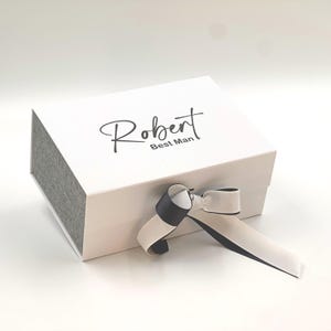 May include: White gift box with a gray lid and a black and white ribbon. The box is personalized with the name "Robert" and the words "Best Man".