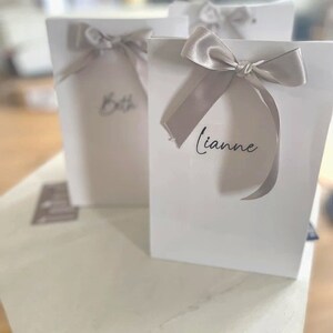 May include: Three white gift bags with silver ribbon bows. The bags are labeled with the names "Beth" and "Lianne".