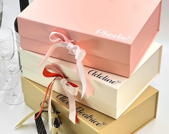 May include: Three gift boxes stacked on top of each other. The top box is pink with the name "Phoebe" written on it. The middle box is white with the name "Adeline" written on it. The bottom box is gold with the name "Alex Beatrice" written on it. Each box has a ribbon tied around it.