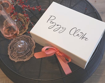 May include: A white rectangular gift box with the name Peggy Clarke printed in black script on the lid, featuring a coral pink ribbon tied in a bow at the front, accompanied by two wine glasses and two brown laser-cut wood coasters.