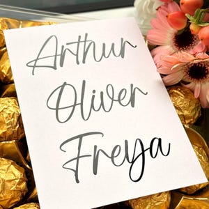 Peut inclure: Une carte blanche avec les noms "Arthur", "Oliver" et "Freya" en écriture grise élégante. La carte est entourée de chocolats dorés et d'un bouquet de fleurs roses et oranges.