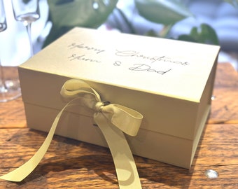 May include: A gold gift box with a yellow ribbon tied in a bow. The box has the words "Merry Christmas Mum & Dad" written on the top.