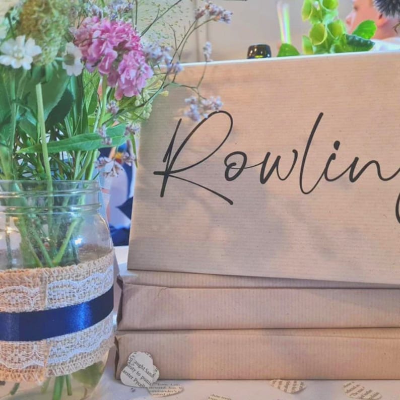 May include: A stack of brown paper-wrapped books with the name "Rowling" written in black cursive on the top book. A vase with a burlap and blue ribbon wrap holds a bouquet of pink and white flowers.
