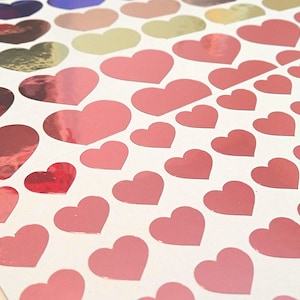 Puede incluir: Una hoja de pegatinas en forma de corazón rosa y dorado. Las pegatinas están dispuestas en filas y todas son del mismo tamaño. Las pegatinas son brillantes y tienen un acabado metálico.