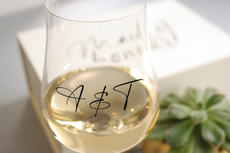 May include: A clear glass wine glass with a gold liquid inside. The glass has the letters "A & T" written on it in black.