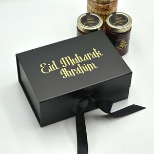 May include: A black gift box with a gold ribbon and the text "Eid Mubarak Ibrahim" on the top. The box is sitting on a white surface and there are three jars of honey in the background.