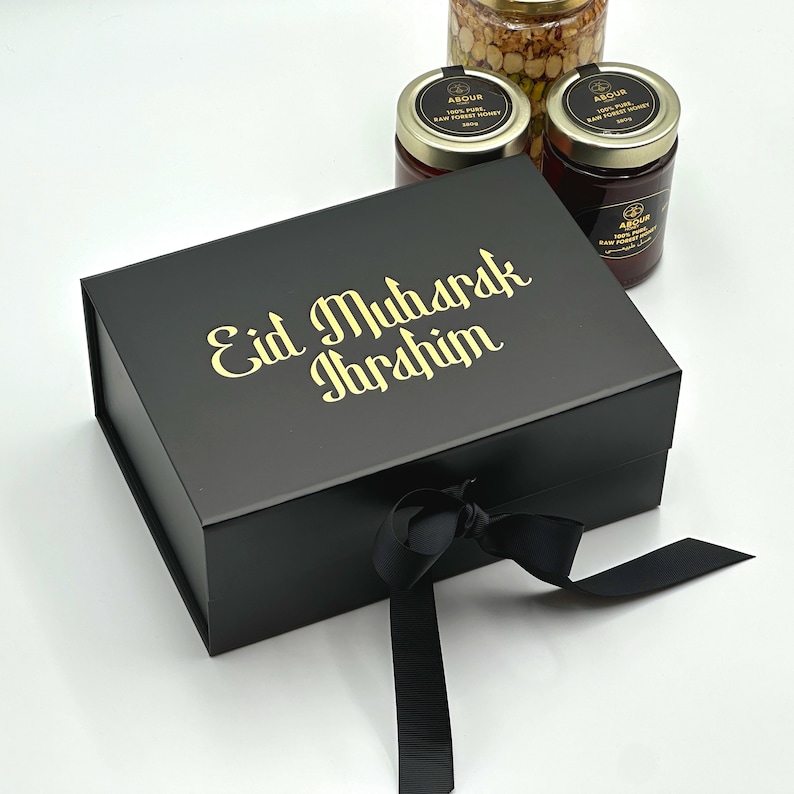 May include: A black gift box with a gold ribbon and the text "Eid Mubarak Ibrahim" on the top. The box is sitting on a white surface and there are three jars of honey in the background.