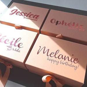 May include: Four pink gift boxes with gold ribbon bows. The boxes are personalized with the names "Jessica bridesmaid", "Ophelie", "Cecile Bride", and "Melanie happy birthday!"