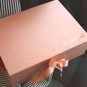 Op de afbeelding: Een lichtroze cadeaudoos met een satijnen lint dat in een strik is gebonden. Op de doos staat in wit handschrift "bridesmaid".