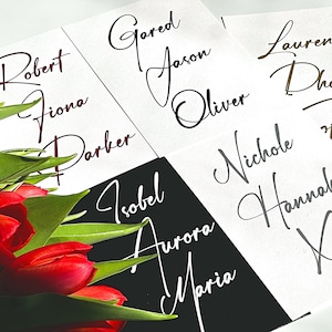 May include: A set of white place cards with black calligraphy names, including Robert, Fiona, Parker, Gared, Jason, Oliver, Lauren,  Nichole, Hannah, and Maria. The place cards are arranged on a black surface with a bouquet of red tulips.