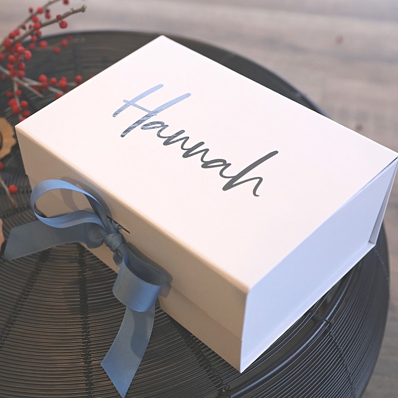 The image shows a white gift box, A5 in size. It has a pastel blue ribbon tied around it. On the top of the lid, the name "Hannah" is written as an example of personalization. The gift box is placed on a round, black metal table. The table.