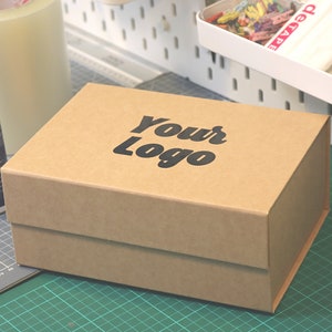 Op de afbeelding: Een bruine kartonnen doos met de tekst "Your Logo" in zwart op het deksel. De doos staat op een groene snijmat, met een rol tape en een lijmfles in de buurt. De doos is waarschijnlijk voor verpakking of opslag.