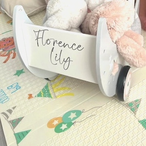 May include: White wooden toy wagon with wheels, featuring a gray moon shape on the side. The wagon has a removable bin with the names "Florence Lily" written on it. The wagon is filled with plush toys, including a white bunny and a pink bunny.