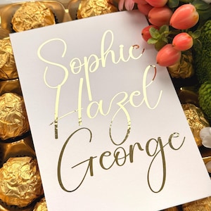 Op de afbeelding: Een witte kaart met gouden cursieve tekst die "Sophie Hazel George" luidt. De kaart is omgeven door goud omwikkelde chocolaatjes en decoratieve bloemen met roze en oranje bessen.