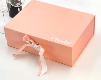 May include: A peach-colored gift box with a white ribbon and bow. The box has the word "Phoebe" printed on the top in white lettering.