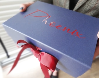 May include: A navy blue gift box with the word "Phoenix" in red script. The box is tied with a red ribbon. The box is held by a person.