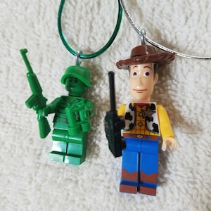 May include: Green Army Man and Woody Lego minifigures on necklaces. The Army Man is holding a rifle and binoculars. Woody is wearing his signature cowboy hat and vest.