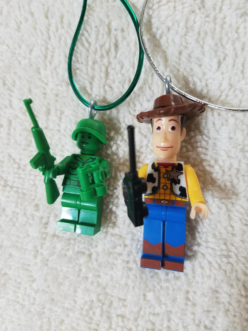 May include: Green Army Man and Woody Lego minifigures on necklaces. The Army Man is holding a rifle and binoculars. Woody is wearing his signature cowboy hat and vest.