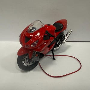 May include: A red toy motorcycle with a black seat and silver exhaust pipes. The motorcycle has a clear windshield and black tires. A red cord is looped in front of the motorcycle.